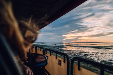 Von einem Autozug heraus fotografiert, kann über die Begrenzung auf das Meer geblickt werden. Die Sonne geht unter und es herrscht Ebbe. 