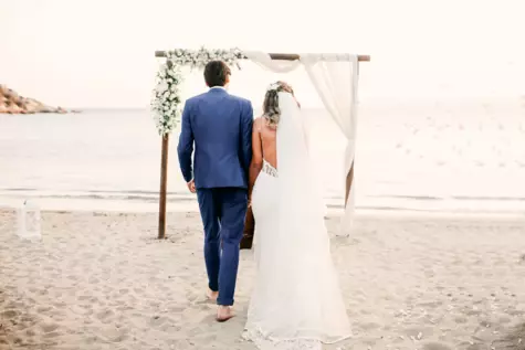 Ein Paar in Anzug und Brautkleid läuft Hand in Hand zu einem dekorierten Bogen am Strand. Im Hintergrund ist das blaue Wasser zu sehen. 