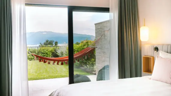 Ein helles elegantes Hotelzimmer mit einem gemütlichen Bett. Eine große Terrassentür trennt den Schlafbereich von einer Terrasse mit einer roten Hängematte und Grünfläche. 