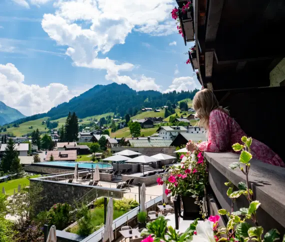 Frau in rosa Kleid lehnt auf dem Balkon des A-ROSA Ifen Hotels und blickt auf den sonnigen Außenbereich mit Pool, Sonnenterrasse und die Berglandschaft des Kleinwalsertals.