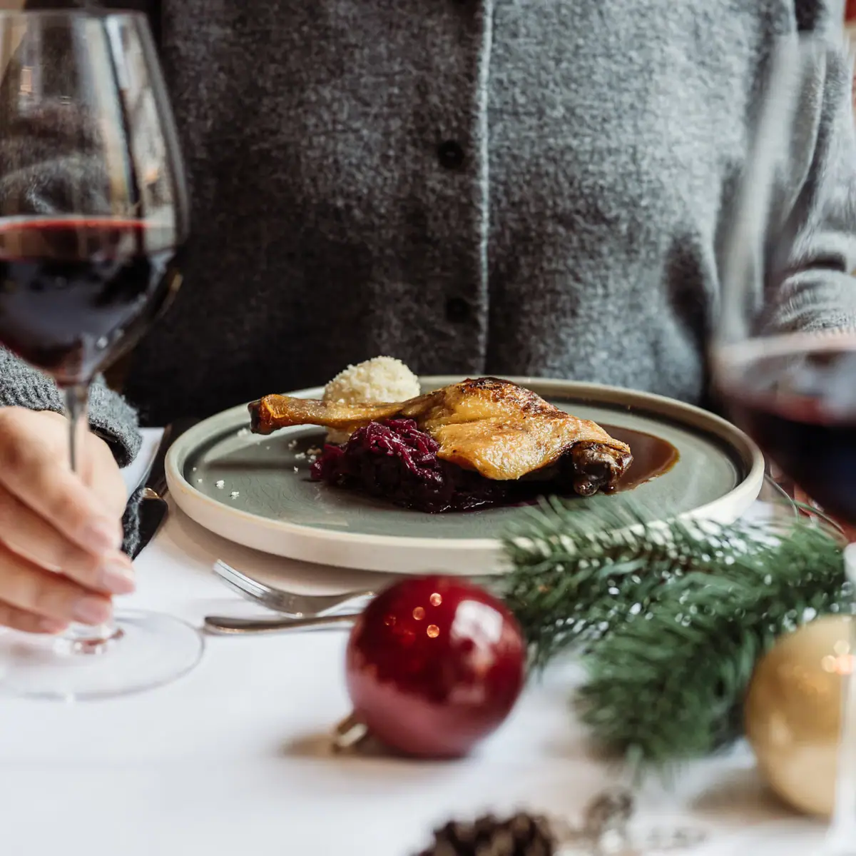 Eine Person isst von einem Teller mit Gänsekeule, Kartoffel und Rotkohl mit weihnachtlichem Tischgedeck und Rotwein im Glas..