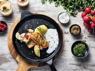 Ein stilvoll angerichtetes Gericht mit gebratenem Fisch, Kartoffeln und Sauce in einer gusseisernen Pfanne, umgeben von frischen Kräutern, Radieschen, Gewürzen und kleinen Dessertgläsern mit Beeren auf einer rustikalen Holzoberfläche.