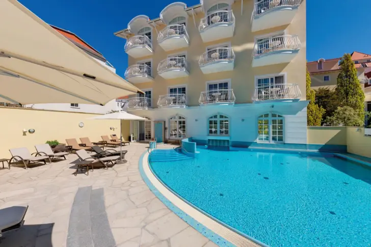 Swimmingpool im Freien umgeben von Stühlen und Sonnenschirmen mit Bäumen und einem klaren Himmel mit untergehender Sonne im Hintergrund.