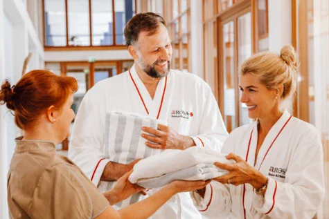 Ein Paar in weißen A-ROSA Bademänteln bekommt weiße Handtücher von einer rothaarigen Mitarbeiterin in beiger Kleidung überreicht.  