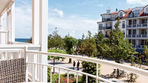 Balkon mit weißem Geländer, mit Blick auf eine Promenade mit Bäumen und einem Gebäude im Hintergrund.