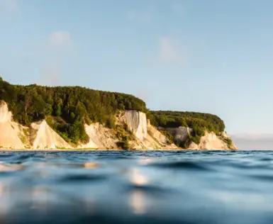 Aus dem blauen, in der Sonne glitzernden Wasser, ragen die hellen, grün bewachsenen Kreidefelsen. 