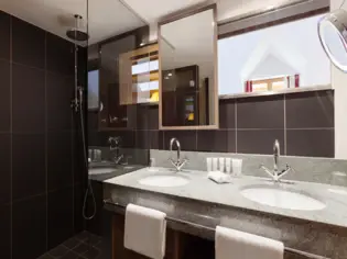 A bathroom with dark tiles, a shower and a large washbasin with two sinks.