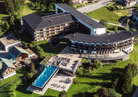 Großes Gebäude umgeben von Grasflächen und Häusern im alpinen Stil, aufgenommen aus der Vogelperspektive.