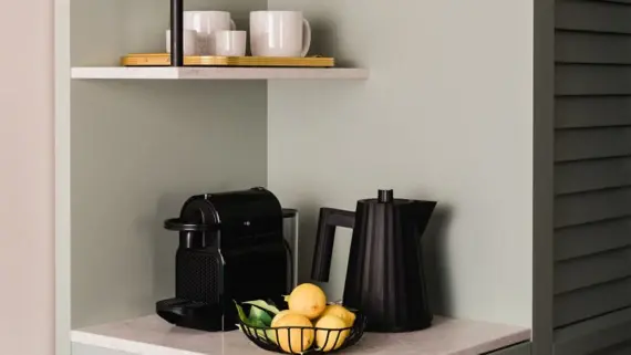 A small, tidy kitchen corner in a superior room with a modern black coffee machine and a stylish kettle on a marble worktop. Above it are two hanging shelves that house some books and two ceramic cups. A basket of fresh lemons provides a splash of color and accentuates the cozy and inviting ambience of the room.