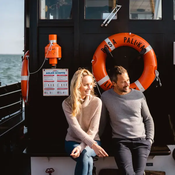 Eine Frau und ein Mann sitzen auf einem kleinen Boot in der Sonne. Hinter Ihnen ist das Kapitänshäuschen mit einem orangenem Rettungsring zu sehen.