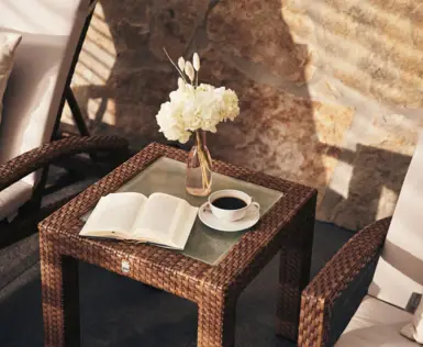 Eine gemütliche Leseecke mit einem geöffneten Buch und einer Tasse Kaffee auf einem Glastisch, umgeben von Rattanmöbeln. Eine Vase mit weißen Blumen und zarten Pampasgräsern ziert den Tisch, was ein elegantes und ruhiges Ambiente schafft. Die Szene spielt sich im sanften Schatten einer Terrasse ab, mit natürlichen Farben und Texturen, die zum Entspannen und Verweilen einladen.