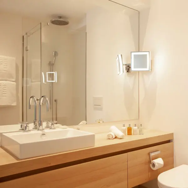 Ein modernes Badezimmer mit einem großem Waschtisch aus hellem Holz, auf dem ein eckiges weißes Waschbecken steht. Im Spiegelbild ist eine verglaste Duschkabine zu sehen.