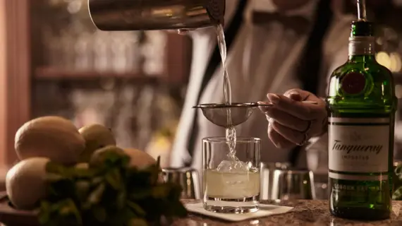 Person gießt Flüssigkeit aus einem Cocktail-Shaker durch ein Sieb in ein Glas.