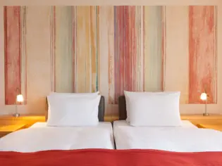 Ein Bett mit vier Kissen und einer roten Tagesdecke steht vor einer orange-roten Tapete.