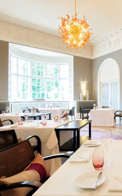 Geräumiges Restaurant mit klassischer Eleganz, großen Fenstern, die reichlich Tageslicht hereinlassen, und kunstvollen Deckenverzierungen. Im Zentrum des Raumes hängt ein moderner orangefarbener Kronleuchter, während an den Wänden zeitgenössische Kunstwerke hängen. Die Tische sind fein mit weißen Tischdecken und roten Gläsern gedeckt, und bieten eine einladende Atmosphäre.