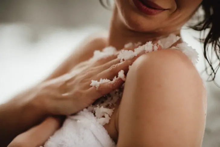 Close-up of a woman applying a salt scrub to her shoulder and arm. The grain of the scrub is clearly visible and emphasizes the care routine. Her relaxed smile suggests a wellness experience. The focus is on the gentle application of the scrub, which conveys a sense of self-care and relaxation.