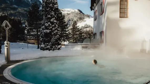 Eine Person schwimmt entspannt im dampfenden Außenpool, umgeben von schneebedeckten Landschaften und einem traditionellen Gebäude. 