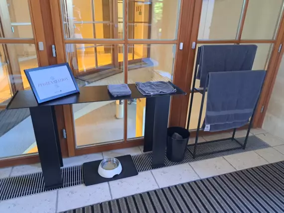 Ein Tisch in einem Hotelbereich mit einem Schild, das "Pfotenstation" anzeigt. Auf dem Tisch liegen gefaltete Handtücher, darunter eine Schale mit Wasser und ein Mülleimer. Im Hintergrund sind große Fenster mit Blick auf einen Innenbereich zu sehen.