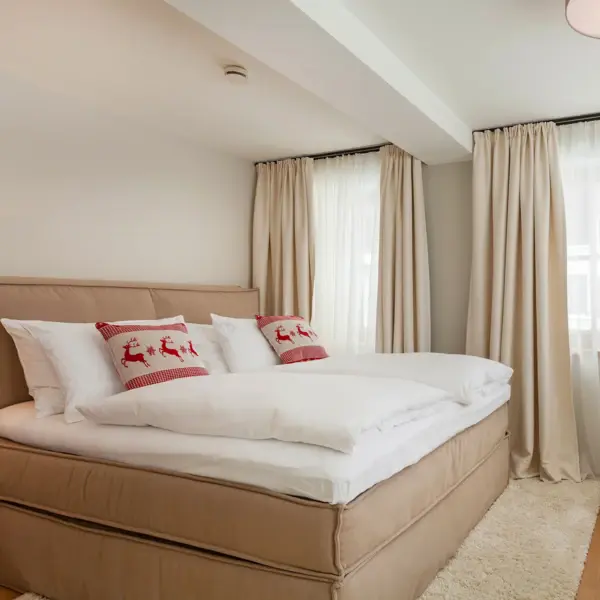 Ein großes gemütliches Polsterbett mit weißer Bettwäsche und zwei roten Deko-Kissen mit Rentieren darauf.