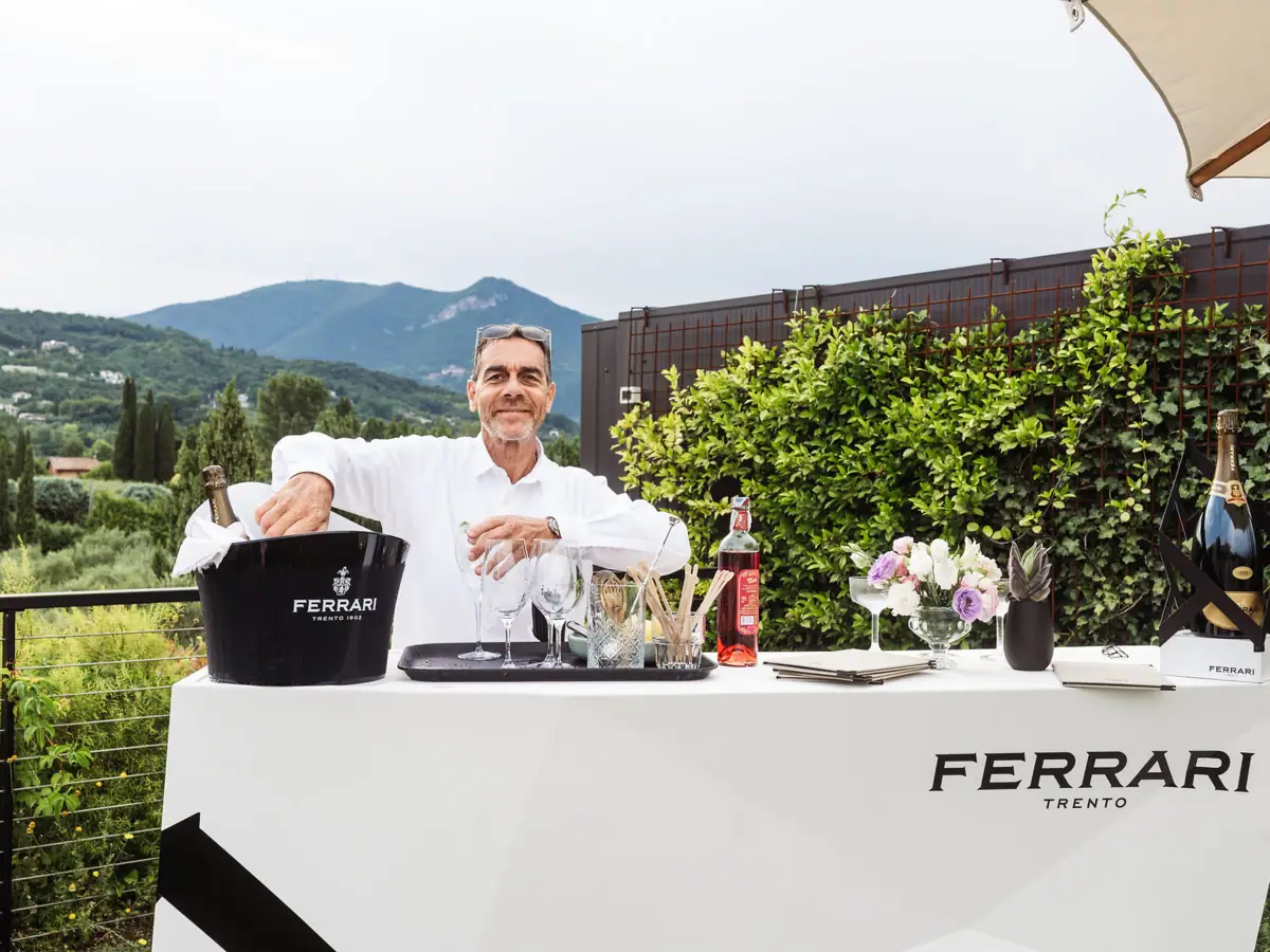 Cocktailkunst im A-ROSA Gardasee Barkeeper bereitet an einem Ferrari-Trento-Tisch Cocktails zu, umgeben von mediterraner Natur und Bergblick.