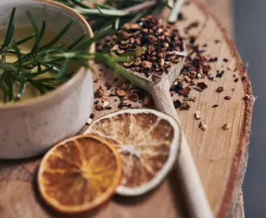 Close-up of a relaxing wellness scene with essential oil and natural ingredients. A small, light-colored ceramic container with clear oil and fresh rosemary sprigs stands on a rustic wooden board. Next to the container are dried orange slices and a wooden spoon full of colorful spices, including pink pepper and dried flower petals scattered across the board, creating an atmosphere of calm and relaxation.