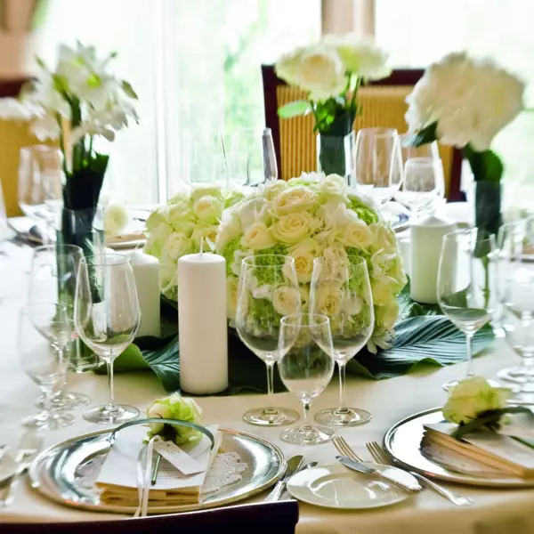 Ein festlich dekorierter Hochzeitstisch mit einem zentralen Blumengesteck aus weißen Rosen und grünen Akzenten. Um das Gesteck sind mehrere große weiße Kerzen gruppiert, die eine warme und einladende Atmosphäre schaffen. Der Tisch ist mit glänzendem Geschirr, Besteck und Gläsern für ein elegantes Essen eingedeckt. Jeder Platz ist mit einer kleinen Rose und einer persönlichen Namenskarte für die Gäste versehen, die auf der weißen Tischwäsche sorgfältig platziert sind. Die natürliche Beleuchtung durch das Fenster betont die Frische und Natürlichkeit der Szenerie.