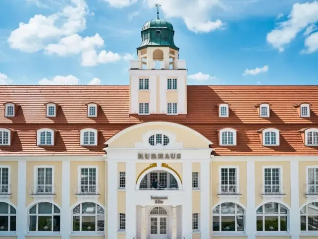 Eine Vorderansicht des hellen Kurhauses binz mit vielen Fenstern und einem roten Dach. 