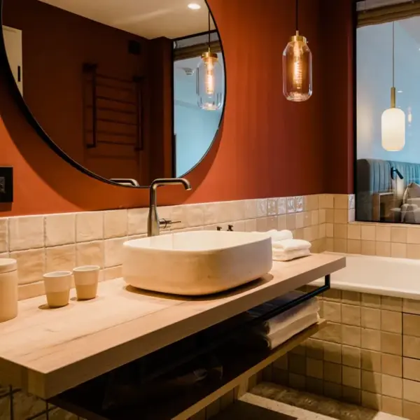 A stylish bathroom with a terracotta-colored wall and beige tiled wash area. A large round mirror hangs above a washbasin with an oval stone washbowl and underneath is an open wooden shelf with towels. Two hanging pendant lights illuminate the room, while the bedroom with a made-up bed is visible through a glass wall in the background.