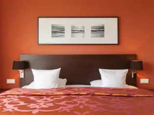 Ein großes Bett mit orangener Tagesdecke steht vor einer orangenen Wand. Das Kopfteil ist aus dunklem Holz und darüber hängt ein länglicher Bilderrahmen mit drei einzelnen quadratischen Motiven.