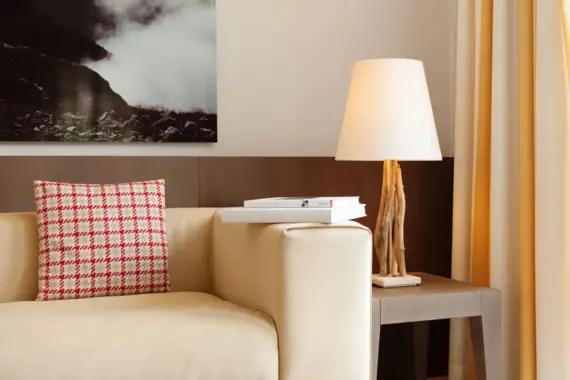 Ein helles Sofa mit einem karierten Kissen steht neben einem Beistelltisch mit einer kleinen Lampe. Auf der Lehne des Sofas liegen zwei Bücher und an der Wand hängt ein dunkles Bild.