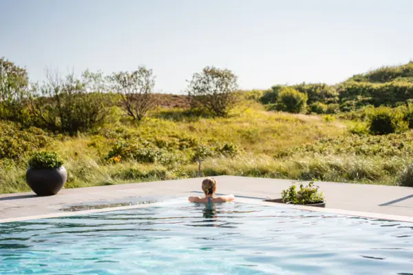Außenpool Sylt Eine Frau lehnt sich an den Beckenrand von einem Außenpool und schaut auf eine Dünenlandschaft.