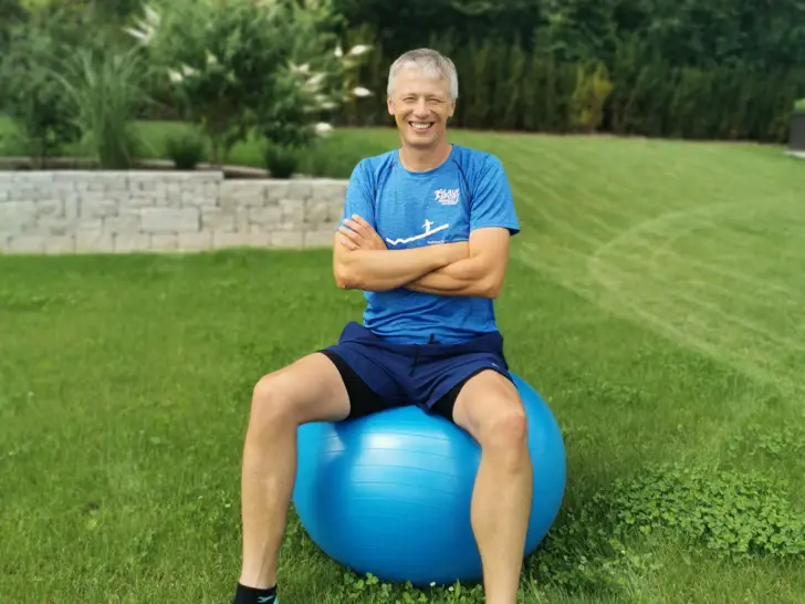 Mann sitzt draußen auf einem großen blauen Gymnastikball auf einer Wiese.