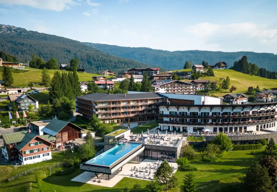 An einem Außenpool von dem A-ROSA Gardasee stehen ordentlich aufgereihte Liegen mit Sonnenschirmen. Im Hintergrund sind Berge und ein Dorf an einem See zu sehen. Die Sonne scheint warm und glitzert auf dem Poolwasser. 