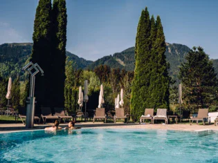 Im Vordergrund steht ein Außenpool in dem zwei Frauen baden. Von hinten zu sehen stützen sie sich auf den Rand des Beckens. Umrandet ist der Pool im Hintergrund von einem Sonn- und Liegebereich mit Liegen und zusammengeklappten Sonnenschirmen. Dahinter stehen hohe Bäume und eine grüne Berglandschaft ist zu sehen.  