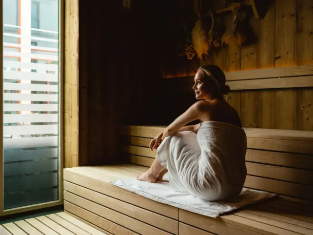  Eine Frau entspannt sich in einer Sauna und blickt sinnierend zur Seite. Sie ist in ein weißes Handtuch gehüllt und sitzt auf einer hölzernen Bank, unter ihr liegt ein weißes Handtuch. Durch die großen Fenster fällt natürliches Licht herein und betont die ruhige und erholsame Atmosphäre der Szene. Die Holzwände der Sauna vermitteln Wärme und Gemütlichkeit.