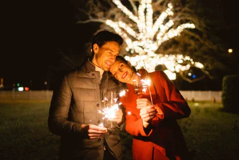 Ein Paar ist lächelnd aneinander gelehnt. Sie stehen warm angezogen bei Nacht draußen und halten Wunderkerzen in ihren Händen. Im Hintergrund leuchtet ein mit Lichterketten verzierter Baum. 