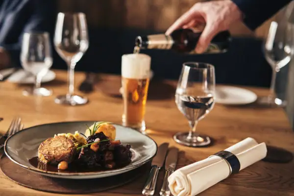 Auf einem gedeckten Holztisch steht schön angerichtetes, deftiges Gericht auf einem Teller, während eine Hand im Hintergrund Bier in ein Glas eingießt.