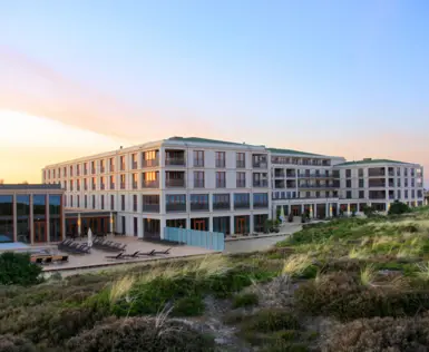 Modernes Hotelgebäude inmitten einer Dünenlandschaft bei Sonnenuntergang. Die Fassade des Hotels spiegelt das warme Licht des Himmels wider, während Liegen und Terrassenbereiche eine entspannte Atmosphäre schaffen.