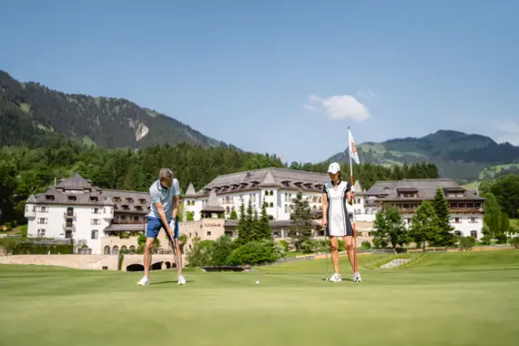 Ein Mann beim Golfspiel auf einem gepflegten Grün, während eine Frau mit einer Fahnenstange daneben steht. Im Hintergrund sind elegante Hotelgebäude und eine malerische Berglandschaft zu sehen.