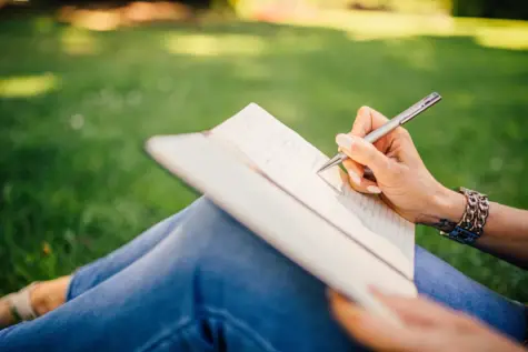 Eine Person sitzt im Gras und schreibt in ein aufgeschlagenes Notizbuch. Man sieht die gekreuzten Beine in Jeans, das Notizbuch auf dem Schoß und eine Hand, die einen Stift hält. Ein Armband und eine Uhr schmücken das Handgelenk. Der Fokus liegt auf der schreibenden Hand, der Hintergrund ist unscharf und lässt grünes Gras erkennen.