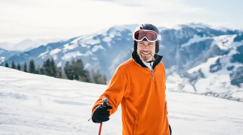 Ein Mann in orangener Skikleidung steht auf einer schneebedeckten Skipiste und lächelt. Im Hintergrund erstreckt sich eine beschneite Berglandschaft, der Himmel ist sonnig und leicht bewölkt. 