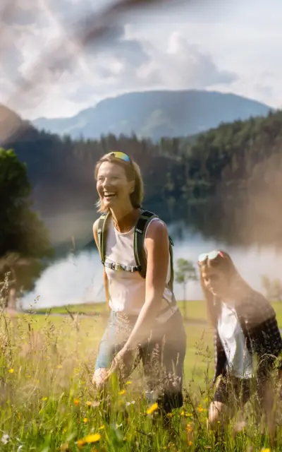 Zwei Frauen wandern in einem Feld, umgeben von Gras und Blumen vor einer Bergkulisse.