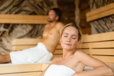 Ein Mann und eine Frau sitzen in Handtücher gewickelt entspannt in einer Holzsauna. 