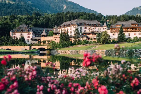 Ein großes, traditionelles Gebäude am Ufer eines Sees, umgeben von bewaldeten Hügeln, mit der aufgehenden Sonne im Hintergrund