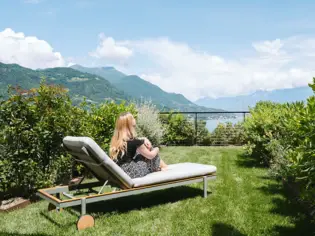 A woman sits with her legs bent on a sun lounger in the green and enjoys the view. The sun is shining warmly. 