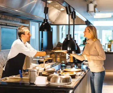 Zwei Frauen treten an eine Live-Cooking Station heran, an der ein Koch in schwarzer Arbeitskleidung Speisen zubereitet. 