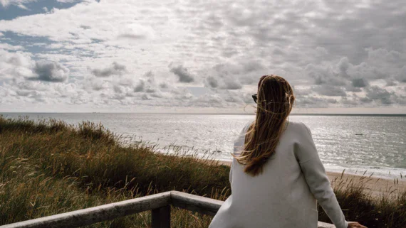Eine Frau blickt auf das Meer hinaus, sie steht am Geländer einer Aussichtsplattform, umgeben von Dünengras. Der Himmel ist mit vielen Wolken bedeckt, die sich über die weite, ruhige See erstrecken. 