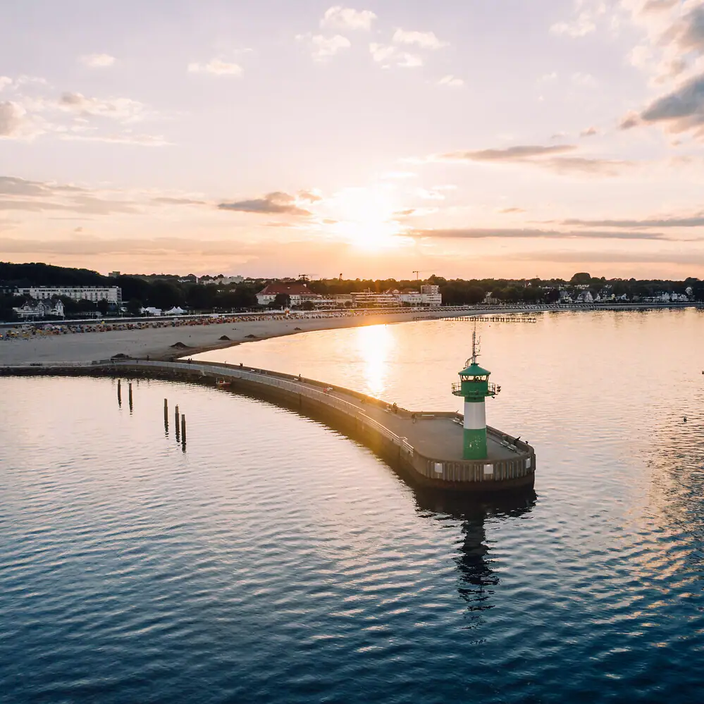 Ein Pier mit einem Leuchtturm darauf, umgeben von Wasser und Wolken am Himmel.