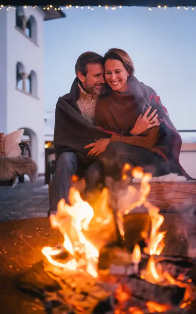 Ein Mann und eine Frau sitzen lächelnd auf einer Bank neben einem Lagerfeuer.