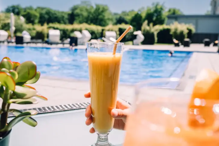 Eine Hand hält einen gelben Smoothie in einem hohen Glas vor einem Außenpool.